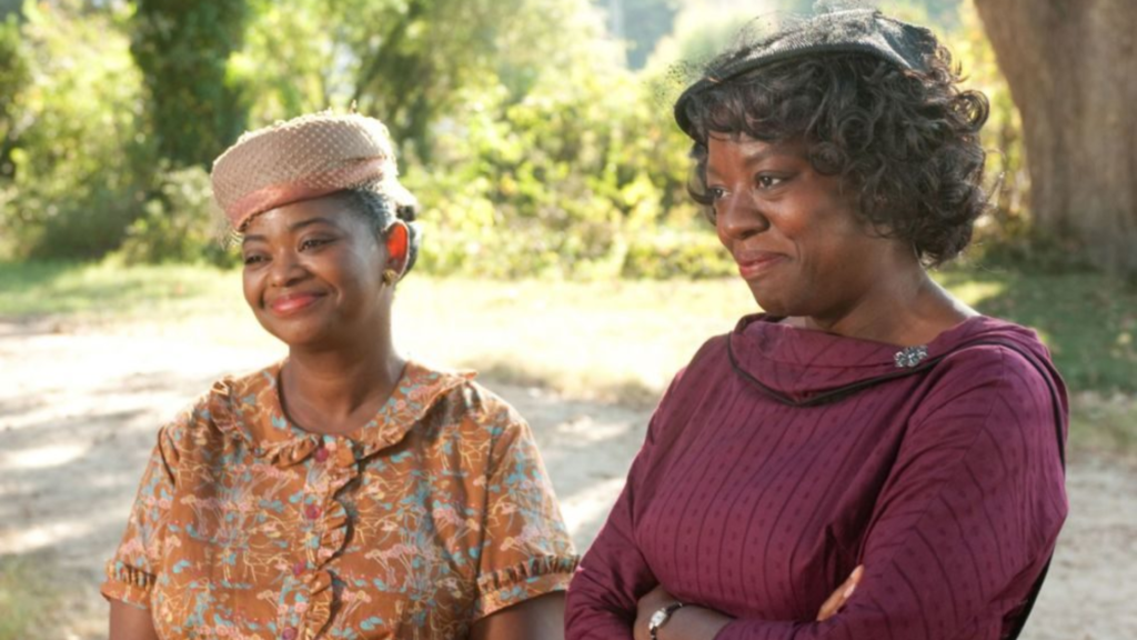 Cena do filme "Histórias Cruzadas" mostrando duas mulheres negras em um ambiente externo ensolarado. À esquerda, Octavia Spencer usa um chapéu rosa claro e um vestido estampado, sorrindo de forma gentil. À direita, Viola Davis usa um chapéu preto e um vestido cor de vinho, com os braços cruzados e uma expressão de orgulho e serenidade.