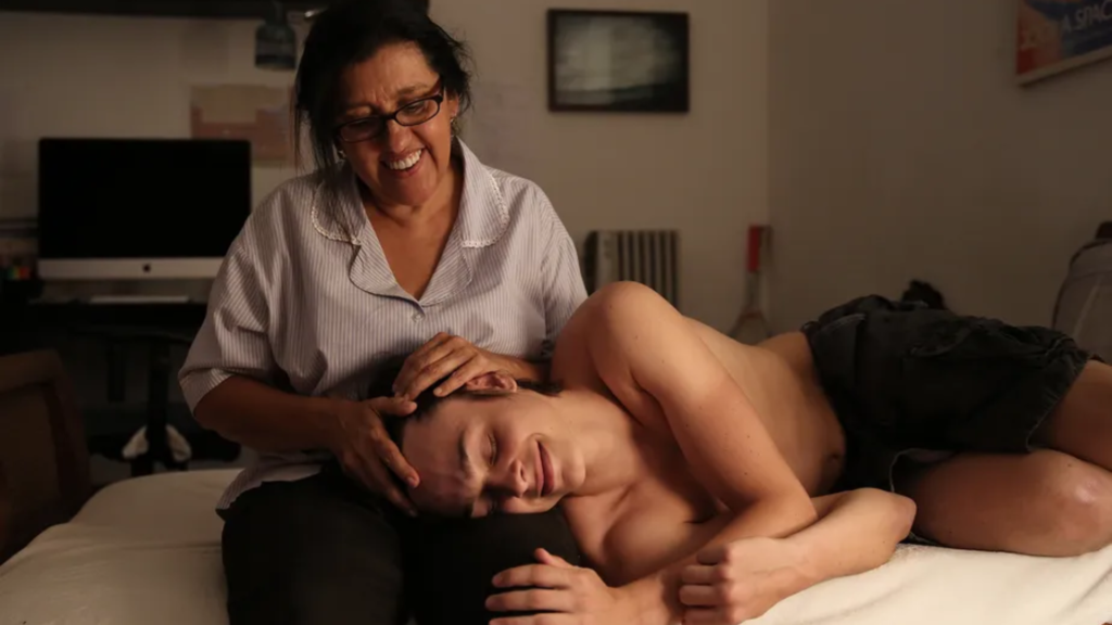 Cena do filme "Que Horas Ela Volta?" em um quarto com iluminação suave. Uma mulher de meia-idade, com óculos e cabelos escuros presos, sorri com ternura enquanto está sentada em uma cama. Em seu colo, um jovem está deitado de lado, com os olhos fechados e uma expressão serena. Ela repousa as mãos carinhosamente sobre a cabeça dele, em um gesto de acolhimento e cuidado.