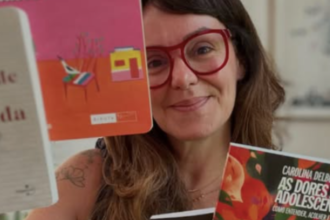 A imagem apresenta a psicanalista e colunista Carolina Delboni, figura central no debate sobre educação e comportamento adolescente mencionado em seu texto. Descrição Detalhada: A figura central: Carolina aparece em primeiro plano, com um semblante acolhedor e um leve sorriso. Ela usa óculos de armação grossa e avermelhada, que se destacam em seu rosto. Elementos visuais: Ela segura e exibe livros, reforçando sua autoridade no tema educacional. No canto inferior direito, destaca-se claramente a capa de um de seus livros, intitulado "As Dores da Adolescência: Como entender, acolher e cuidar". Composição: A foto tem um estilo intimista e intelectual. O fundo está levemente desfocado, o que direciona toda a atenção para ela e para as obras que simbolizam seu trabalho de orientação para pais e educadores. Simbolismo: A imagem comunica proximidade e cuidado, características que ela defende em seus artigos ao sugerir que a educação dos meninos deve passar pelo acolhimento emocional e pela escuta ativa.