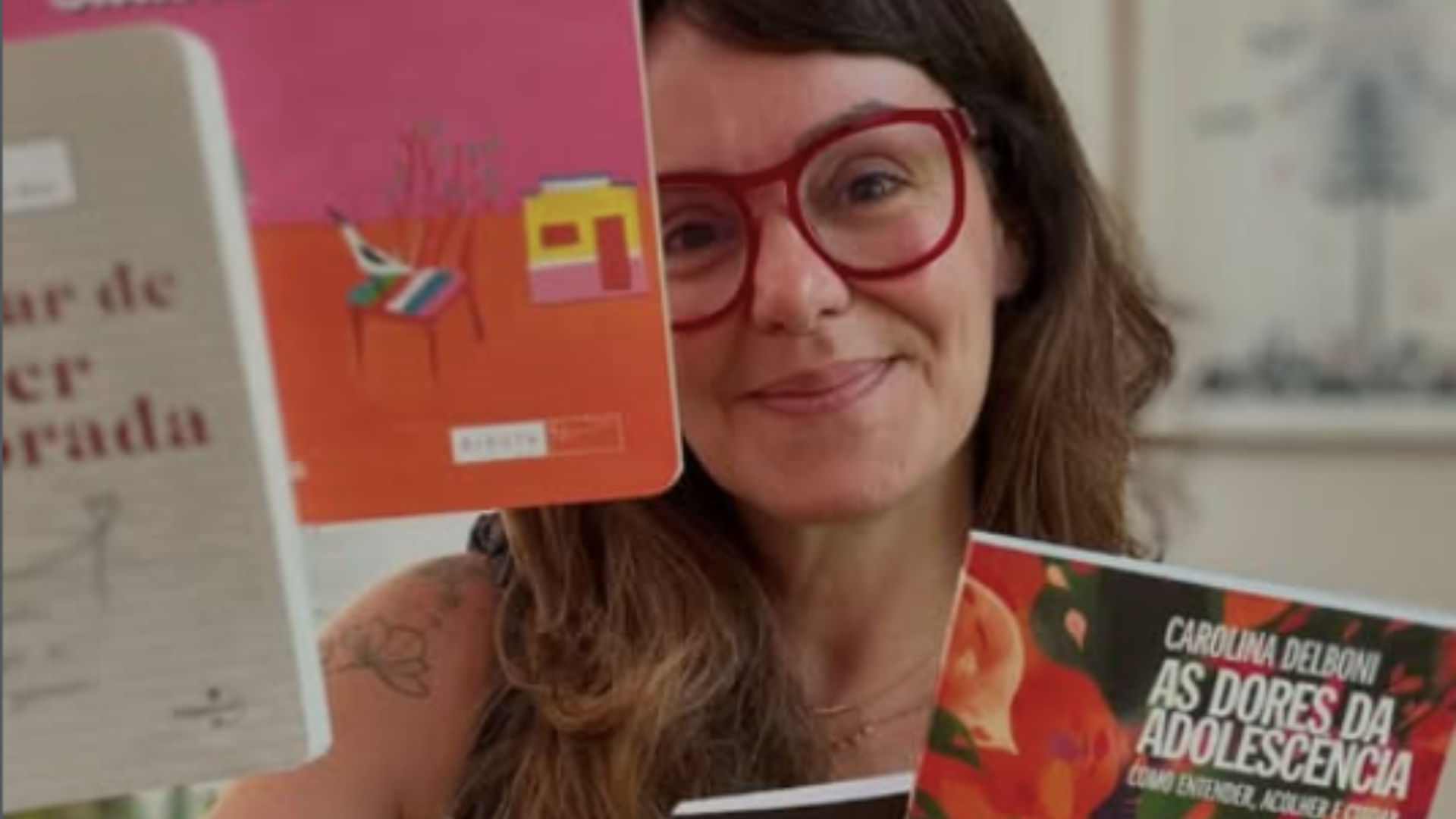 A imagem apresenta a psicanalista e colunista Carolina Delboni, figura central no debate sobre educação e comportamento adolescente mencionado em seu texto. Descrição Detalhada: A figura central: Carolina aparece em primeiro plano, com um semblante acolhedor e um leve sorriso. Ela usa óculos de armação grossa e avermelhada, que se destacam em seu rosto. Elementos visuais: Ela segura e exibe livros, reforçando sua autoridade no tema educacional. No canto inferior direito, destaca-se claramente a capa de um de seus livros, intitulado "As Dores da Adolescência: Como entender, acolher e cuidar". Composição: A foto tem um estilo intimista e intelectual. O fundo está levemente desfocado, o que direciona toda a atenção para ela e para as obras que simbolizam seu trabalho de orientação para pais e educadores. Simbolismo: A imagem comunica proximidade e cuidado, características que ela defende em seus artigos ao sugerir que a educação dos meninos deve passar pelo acolhimento emocional e pela escuta ativa.