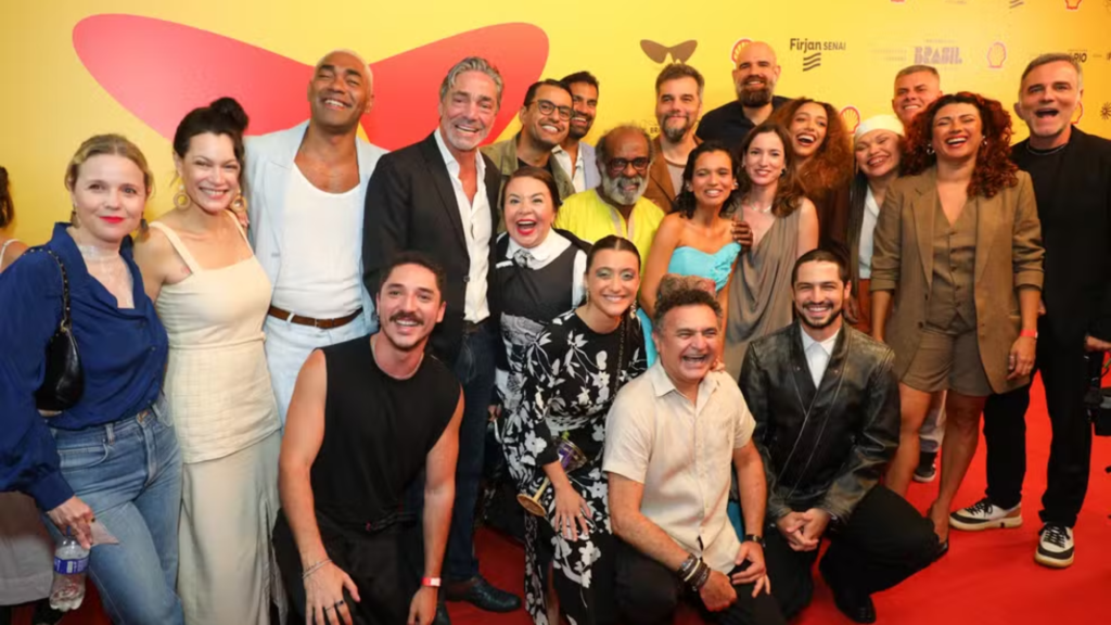 Uma fotografia em grupo reúne o numeroso elenco e a equipe do filme "O Agente Secreto" em um evento formal. Cerca de vinte pessoas posam sorridentes e abraçadas sobre um tapete vermelho, tendo ao fundo um painel amarelo vibrante com logotipos de patrocinadores e uma grande ilustração abstrata em vermelho que lembra as asas de uma borboleta.

O grupo apresenta uma composição diversa de idades e estilos: homens e mulheres vestem trajes que variam entre vestidos elegantes, ternos casuais e roupas modernas sem manga. No centro da imagem, destacam-se figuras como o ator Wagner Moura e a atriz Maria Fernanda Cândido, cercados por outros integrantes do elenco que aparecem tanto de pé quanto agachados à frente, transmitindo uma atmosfera de celebração e união.