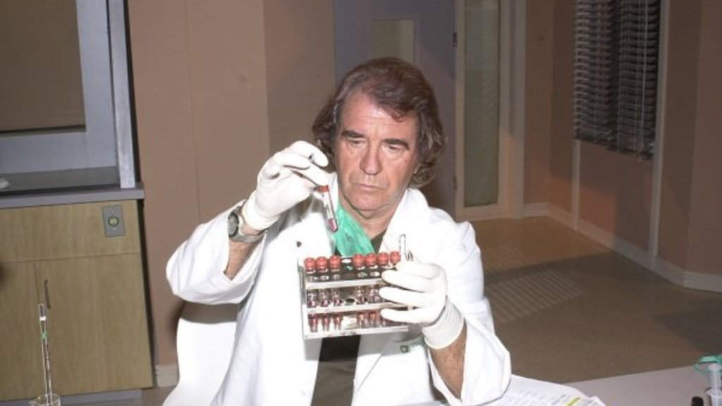 O ator Juca de Oliveira caracterizado como o personagem Dr. Albieri, da novela "O Clone". Ele aparece sentado em um laboratório, vestindo um jaleco branco e luvas descartáveis claras. Com uma expressão concentrada, ele segura um tubo de ensaio com líquido avermelhado em uma mão e uma estante com outros tubos na outra. O ator tem cabelos castanhos ondulados, na altura das orelhas, e o ambiente ao fundo sugere um consultório médico ou laboratório de pesquisa.