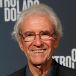 Fotografia de meio-corpo do ator Juca de Oliveira. Ele era um homem idoso, com pele clara, cabelos brancos e curtos, levemente desalinhados. Ele sorri para a câmera e usa óculos de grau com armação fina e escura. Veste um paletó preto sobre uma camisa azul-escura. Ao fundo, uma parede azul-marinho exibe o logotipo da novela "O Outro Lado do Paraíso" em letras brancas.