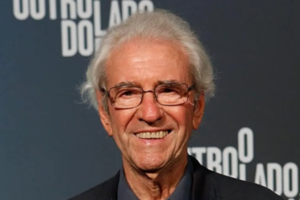 Fotografia de meio-corpo do ator Juca de Oliveira. Ele era um homem idoso, com pele clara, cabelos brancos e curtos, levemente desalinhados. Ele sorri para a câmera e usa óculos de grau com armação fina e escura. Veste um paletó preto sobre uma camisa azul-escura. Ao fundo, uma parede azul-marinho exibe o logotipo da novela "O Outro Lado do Paraíso" em letras brancas.