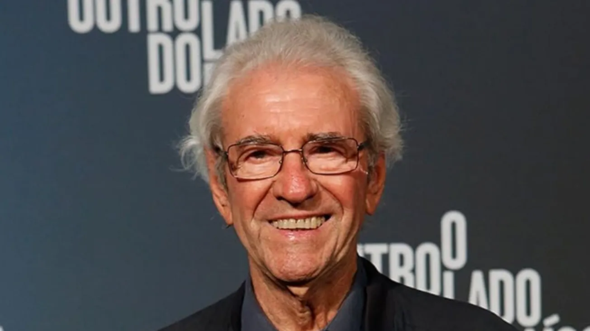 Fotografia de meio-corpo do ator Juca de Oliveira. Ele era um homem idoso, com pele clara, cabelos brancos e curtos, levemente desalinhados. Ele sorri para a câmera e usa óculos de grau com armação fina e escura. Veste um paletó preto sobre uma camisa azul-escura. Ao fundo, uma parede azul-marinho exibe o logotipo da novela "O Outro Lado do Paraíso" em letras brancas.