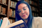 Uma moça de cabelos pretos e óculos de aros grandes lê o livro "O Corcunda de Notre-Dame" sentada em uma biblioteca, com a estante cheia de livros.