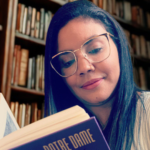 Uma moça de cabelos pretos e óculos de aros grandes lê o livro "O Corcunda de Notre-Dame" sentada em uma biblioteca, com a estante cheia de livros.