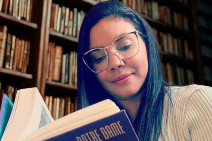 Uma moça de cabelos pretos e óculos de aros grandes lê o livro "O Corcunda de Notre-Dame" sentada em uma biblioteca, com a estante cheia de livros.