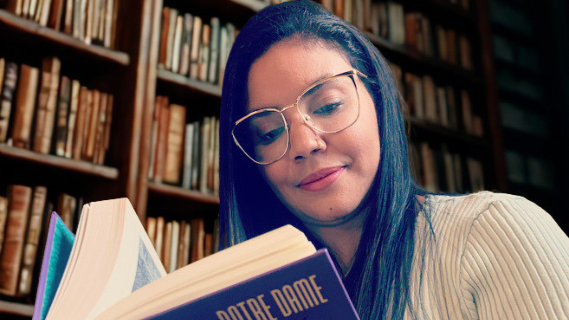 Uma moça de cabelos pretos e óculos de aros grandes lê o livro "O Corcunda de Notre-Dame" sentada em uma biblioteca, com a estante cheia de livros.