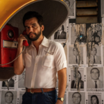 Uma fotografia em plano médio mostra o ator Wagner Moura, barbudo e com cabelo escuro, vestido com uma camisa de botão branca de manga curta, cinto de couro marrom e jeans. Ele está de pé em um orelhão, segurando o fone vermelho do telefone na orelha esquerda com a mão esquerda e olhando para a direita. O orelhão é coberto por uma cúpula amarela. O fundo é uma parede coberta por pôsteres em preto e branco de homens com textos sobrepostos em português.