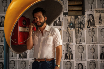 Uma fotografia em plano médio mostra o ator Wagner Moura, barbudo e com cabelo escuro, vestido com uma camisa de botão branca de manga curta, cinto de couro marrom e jeans. Ele está de pé em um orelhão, segurando o fone vermelho do telefone na orelha esquerda com a mão esquerda e olhando para a direita. O orelhão é coberto por uma cúpula amarela. O fundo é uma parede coberta por pôsteres em preto e branco de homens com textos sobrepostos em português.