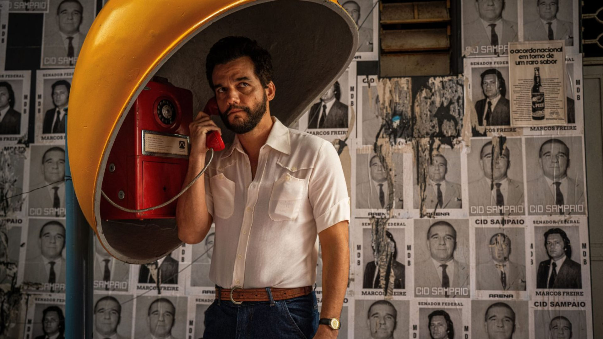 Uma fotografia em plano médio mostra o ator Wagner Moura, barbudo e com cabelo escuro, vestido com uma camisa de botão branca de manga curta, cinto de couro marrom e jeans. Ele está de pé em um orelhão, segurando o fone vermelho do telefone na orelha esquerda com a mão esquerda e olhando para a direita. O orelhão é coberto por uma cúpula amarela. O fundo é uma parede coberta por pôsteres em preto e branco de homens com textos sobrepostos em português.