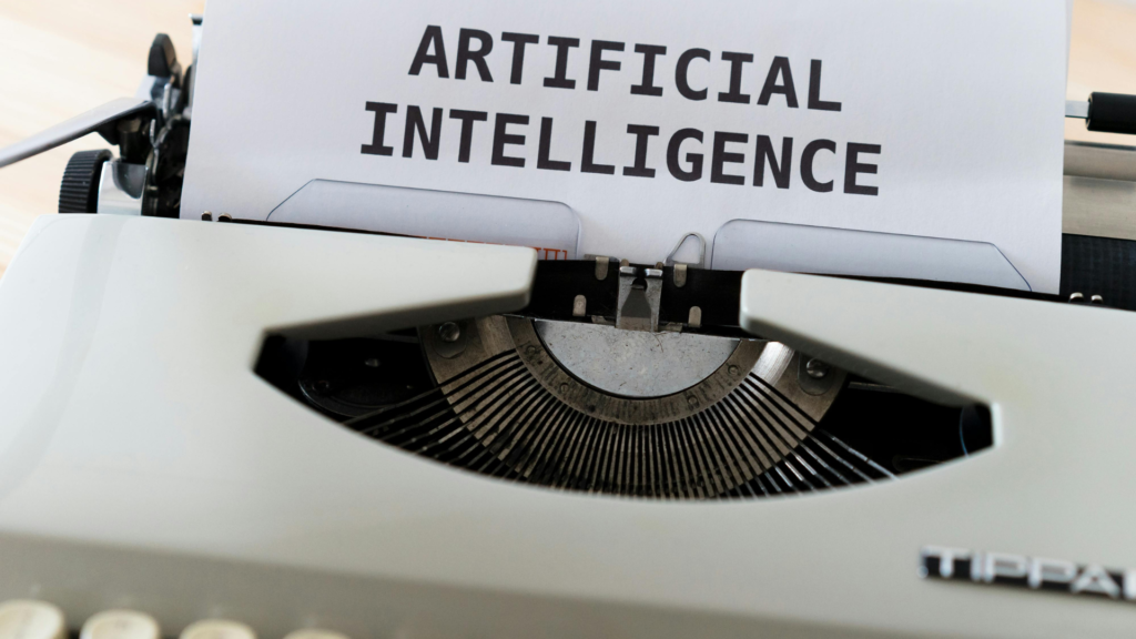 Vista em detalhe e de ângulo superior de uma máquina de escrever vintage de cor cinza-clara. Um papel branco está inserido no rolo da máquina, exibindo as palavras "ARTIFICIAL INTELLIGENCE" impressas em letras maiúsculas, pretas e sem serifa. Parte do teclado com teclas redondas e amareladas aparece desfocada na parte inferior. O foco principal está no mecanismo de tipos e no texto impresso no papel.