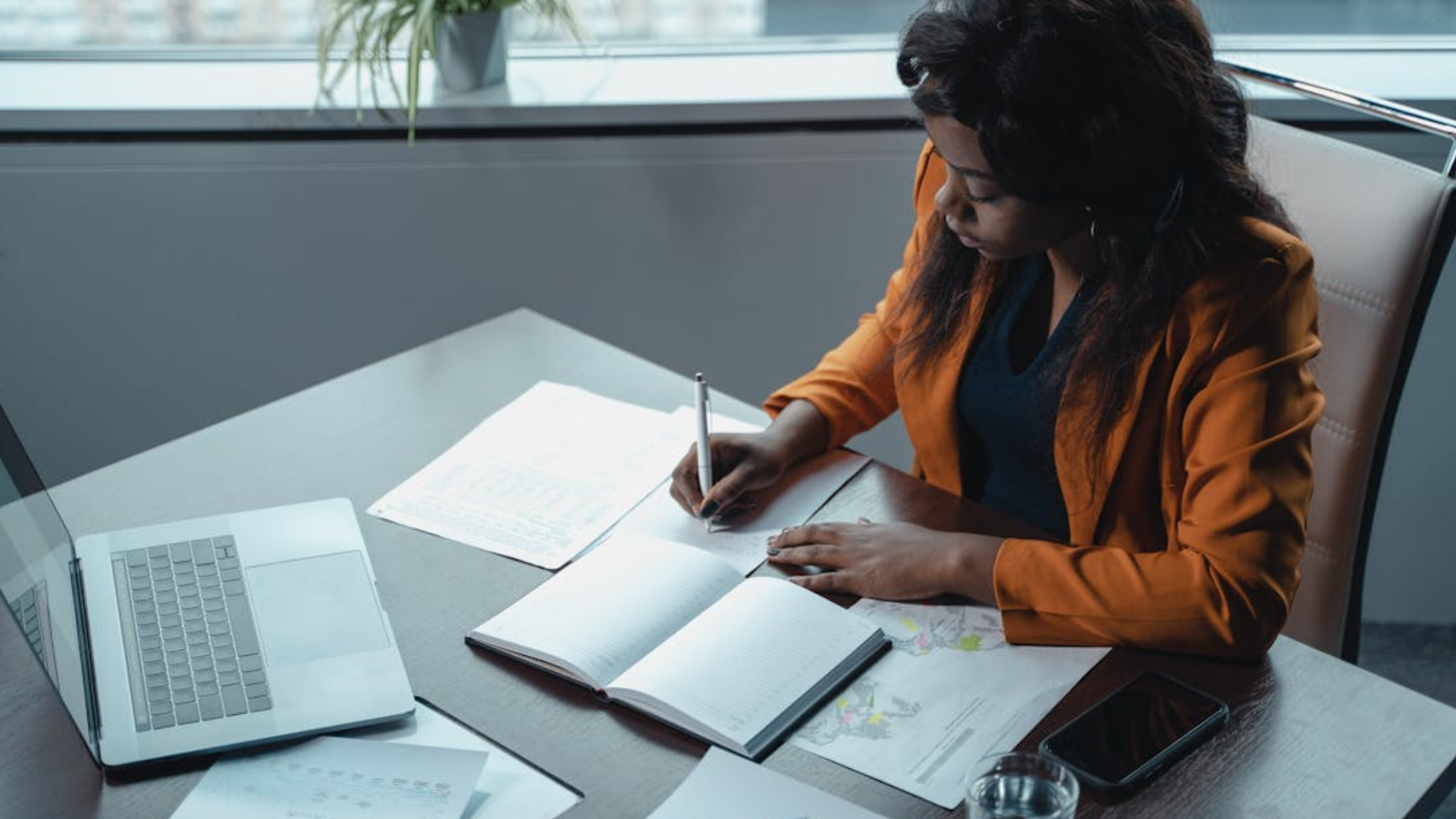 Uma mulher negra com uma jaqueta laranja está sentada em uma mesa, escrevendo em um caderno com uma caneta. Há um notebook aberto ao lado dela, bem como vários papéis e documentos, incluindo gráficos e relatórios. Ela está focada em seu trabalho e tem um copo de água e um smartphone na mesa. Há um peitoril da janela com uma planta atrás dela. A sala está bem iluminada.
