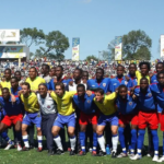 Jogadores das seleções do Brasil e do Haiti posam juntos para foto antes do amistoso conhecido como "Jogo da Paz", realizado em Porto Príncipe em 2004. Ao fundo, o estádio Sylvio Cator aparece lotado de torcedores.