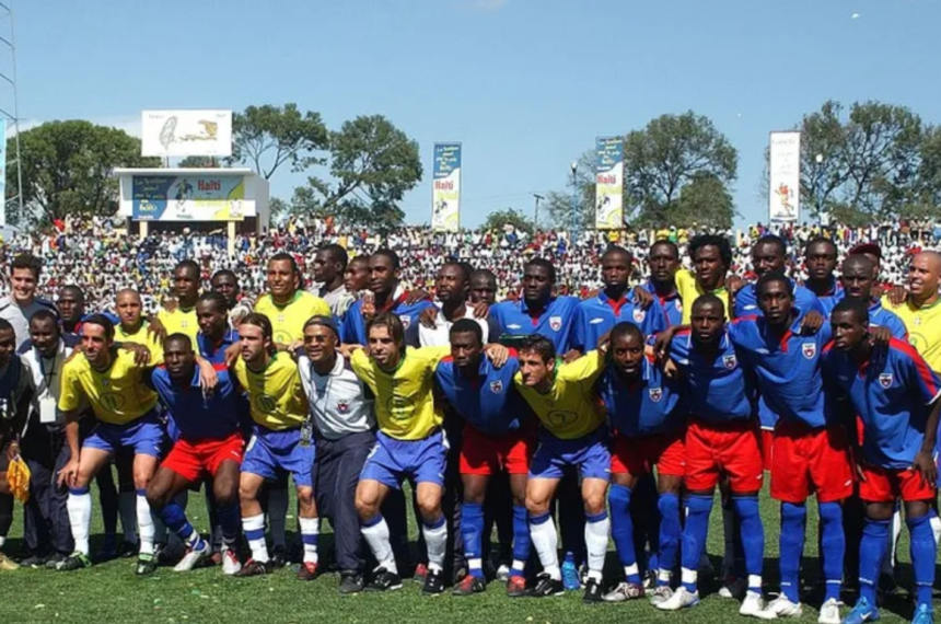 Jogadores das seleções do Brasil e do Haiti posam juntos para foto antes do amistoso conhecido como "Jogo da Paz", realizado em Porto Príncipe em 2004. Ao fundo, o estádio Sylvio Cator aparece lotado de torcedores.