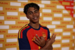Foto de Lamine Yamal, sorrindo, com uniforme da seleção espanhola (vermelho e azul-marinho), em frente a um painel amarelo com o nome "Spain" repetido, fazendo um gesto com as duas mãos próximo ao peito, em uma sessão de fotos oficial da Copa do Mundo.