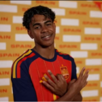 Foto de Lamine Yamal, sorrindo, com uniforme da seleção espanhola (vermelho e azul-marinho), em frente a um painel amarelo com o nome "Spain" repetido, fazendo um gesto com as duas mãos próximo ao peito, em uma sessão de fotos oficial da Copa do Mundo.