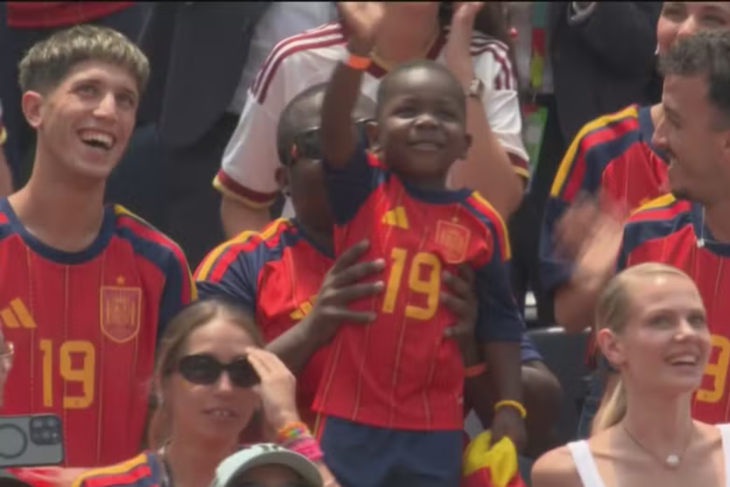 Irmão de Lamine Yamal, com seus três anos, sorridente e carismático, vestindo camisa da seleção espanhola com o número 19, sendo segurada no colo em meio à torcida nas arquibancadas, cercada por outros torcedores sorrindo e comemorando durante uma partida.