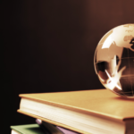 Um globo de vidro transparente equilibrado sobre uma pilha de livros com capas coloridas (dourada, verde e rosa), contra um fundo escuro. A iluminação em tons quentes destaca os continentes gravados no globo e as bordas douradas das páginas dos livros.