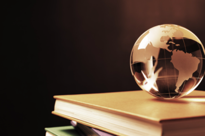 Um globo de vidro transparente equilibrado sobre uma pilha de livros com capas coloridas (dourada, verde e rosa), contra um fundo escuro. A iluminação em tons quentes destaca os continentes gravados no globo e as bordas douradas das páginas dos livros.