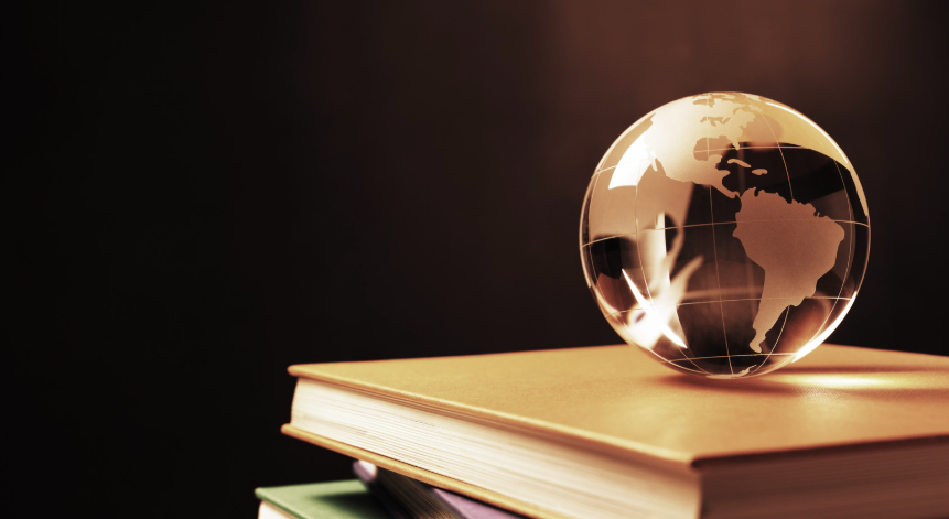 Um globo de vidro transparente equilibrado sobre uma pilha de livros com capas coloridas (dourada, verde e rosa), contra um fundo escuro. A iluminação em tons quentes destaca os continentes gravados no globo e as bordas douradas das páginas dos livros.