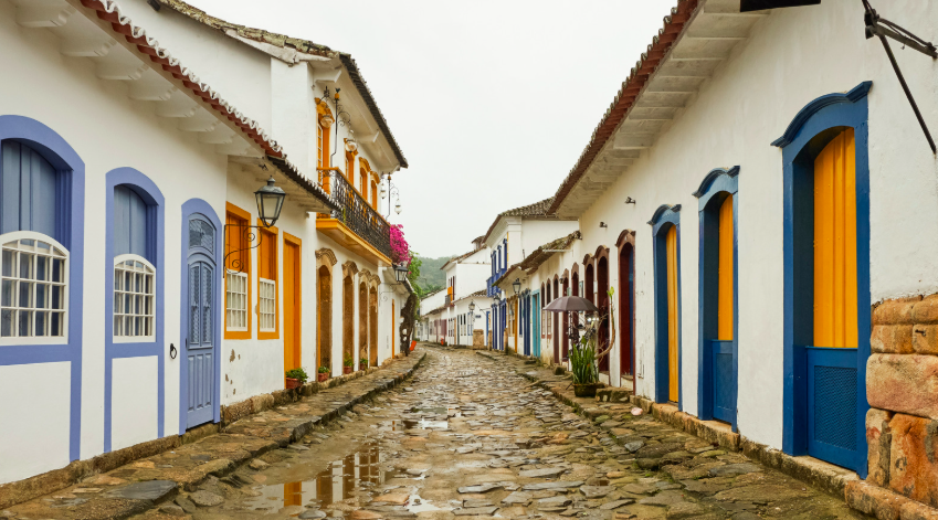 Rua de pedras do centro histórico de Paraty (RJ) ladeada por casarões coloniais brancos com portas e janelas coloridas em azul e amarelo.