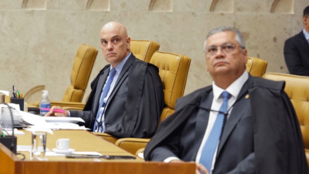Alexandre de Moraes (esquerda) e Flávio Dino (direita) sentam-se à mesa em frente ao Supremo Tribunal Federal (STF) do Brasil. Ambos vestem vestes pretas soltas semelhantes a batas, usadas por juízes e advogados em tribunais brasileiros. Ambos estão sentados em cadeiras de couro dourado. O homem da esquerda olha para a direita e para a frente enquanto o homem da direita olha apenas para a frente. O homem da esquerda tem sua mão direita na mesa ao lado de uma caneta rosa e branca. Há uma garrafa de desinfetante para as mãos e outros itens ao lado da mão dele. Ambos têm papéis na mesa à sua frente e o homem da direita tem uma xícara de chá ou café. Suas gravatas são brancas com linhas azuis. O homem da direita tem um colar na vestimenta. Um homem com paletó e gravata pretas está sentado parcialmente à direita dos dois homens sentados na cadeira do fundo, olhando para frente. Atrás deles, há uma parede de pedra calcária texturizada.