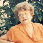 Foto de Sylvia Orthof, mulher de cabelos claros e cacheados, óculos de armação clara, vestindo blusa laranja, sentada ao ar livre com vegetação verde ao fundo.