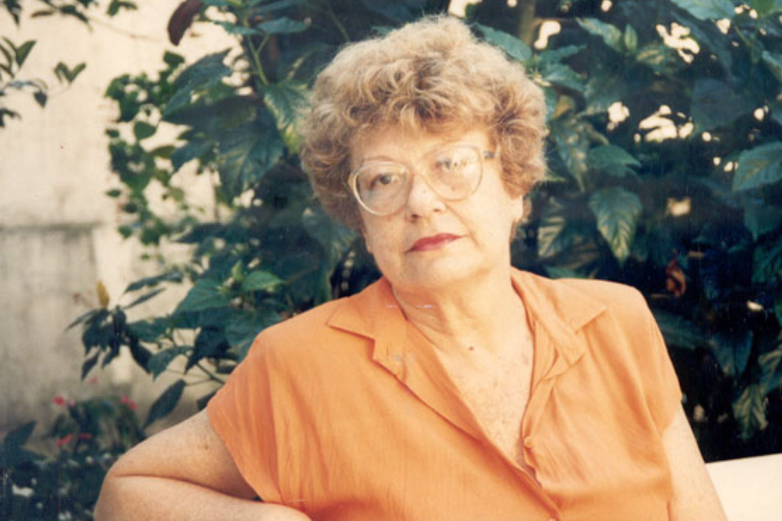 Foto de Sylvia Orthof, mulher de cabelos claros e cacheados, óculos de armação clara, vestindo blusa laranja, sentada ao ar livre com vegetação verde ao fundo.