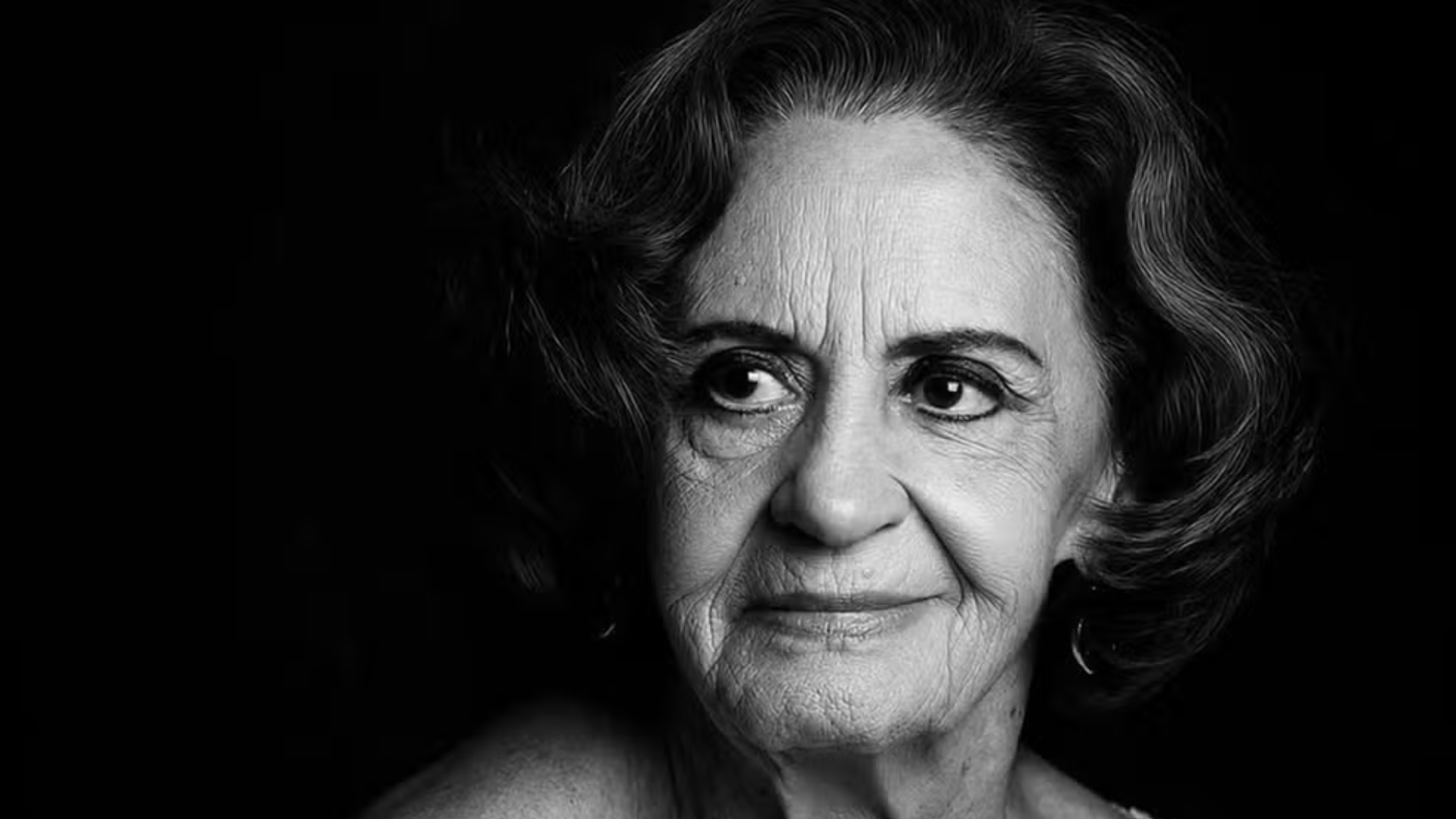 Retrato em preto e branco da atriz brasileira Laura Cardoso, com iluminação dramática vinda da esquerda, destacando as expressões faciais e marcas de idade. Ela olha para o lado com um leve sorriso. O fundo é totalmente escuro.