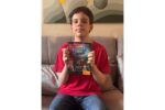 Um menino, vestindo uma camiseta vermelha e calça jeans, está sentado em um sofá cinza, segurando um livro intitulado "Elemento H" voltado para a câmera. A capa do livro apresenta uma ilustração com várias figuras de desenho animado e texto que diz "Elemento H", "José Murilo Bernal Barreto Carneiro" e "A vida na Tabela Periódica". O plano de fundo é uma parede com uma grande pintura de formas geométricas em tons de cinza e amarelo. A foto é tirada de frente, capturando o menino da cintura para cima, e a iluminação é suave e natural.