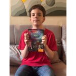 Um menino, vestindo uma camiseta vermelha e calça jeans, está sentado em um sofá cinza, segurando um livro intitulado "Elemento H" voltado para a câmera. A capa do livro apresenta uma ilustração com várias figuras de desenho animado e texto que diz "Elemento H", "José Murilo Bernal Barreto Carneiro" e "A vida na Tabela Periódica". O plano de fundo é uma parede com uma grande pintura de formas geométricas em tons de cinza e amarelo. A foto é tirada de frente, capturando o menino da cintura para cima, e a iluminação é suave e natural.