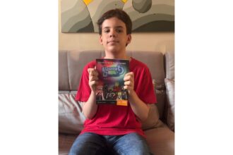 Um menino, vestindo uma camiseta vermelha e calça jeans, está sentado em um sofá cinza, segurando um livro intitulado "Elemento H" voltado para a câmera. A capa do livro apresenta uma ilustração com várias figuras de desenho animado e texto que diz "Elemento H", "José Murilo Bernal Barreto Carneiro" e "A vida na Tabela Periódica". O plano de fundo é uma parede com uma grande pintura de formas geométricas em tons de cinza e amarelo. A foto é tirada de frente, capturando o menino da cintura para cima, e a iluminação é suave e natural.