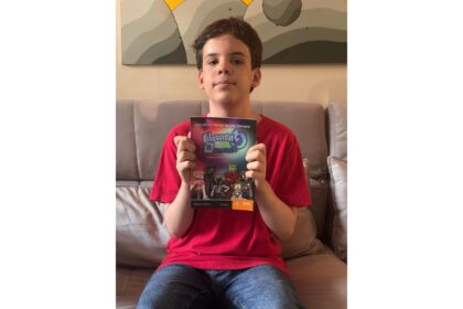 Um menino, vestindo uma camiseta vermelha e calça jeans, está sentado em um sofá cinza, segurando um livro intitulado "Elemento H" voltado para a câmera. A capa do livro apresenta uma ilustração com várias figuras de desenho animado e texto que diz "Elemento H", "José Murilo Bernal Barreto Carneiro" e "A vida na Tabela Periódica". O plano de fundo é uma parede com uma grande pintura de formas geométricas em tons de cinza e amarelo. A foto é tirada de frente, capturando o menino da cintura para cima, e a iluminação é suave e natural.