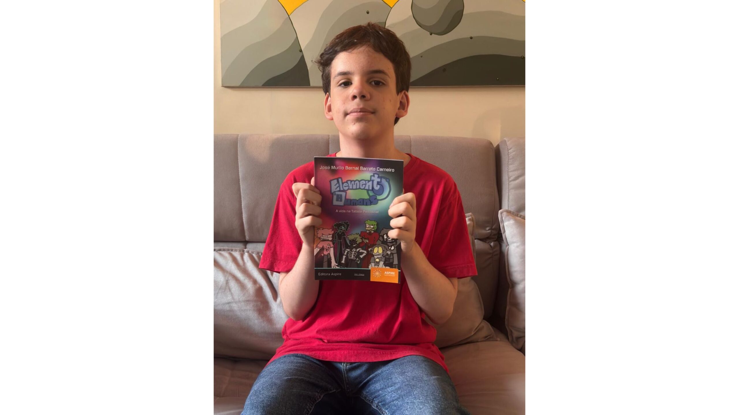 Um menino, vestindo uma camiseta vermelha e calça jeans, está sentado em um sofá cinza, segurando um livro intitulado "Elemento H" voltado para a câmera. A capa do livro apresenta uma ilustração com várias figuras de desenho animado e texto que diz "Elemento H", "José Murilo Bernal Barreto Carneiro" e "A vida na Tabela Periódica". O plano de fundo é uma parede com uma grande pintura de formas geométricas em tons de cinza e amarelo. A foto é tirada de frente, capturando o menino da cintura para cima, e a iluminação é suave e natural.