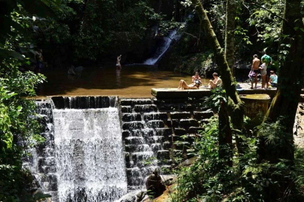 Cachoeira em área de mata, com pessoas à margem da água.