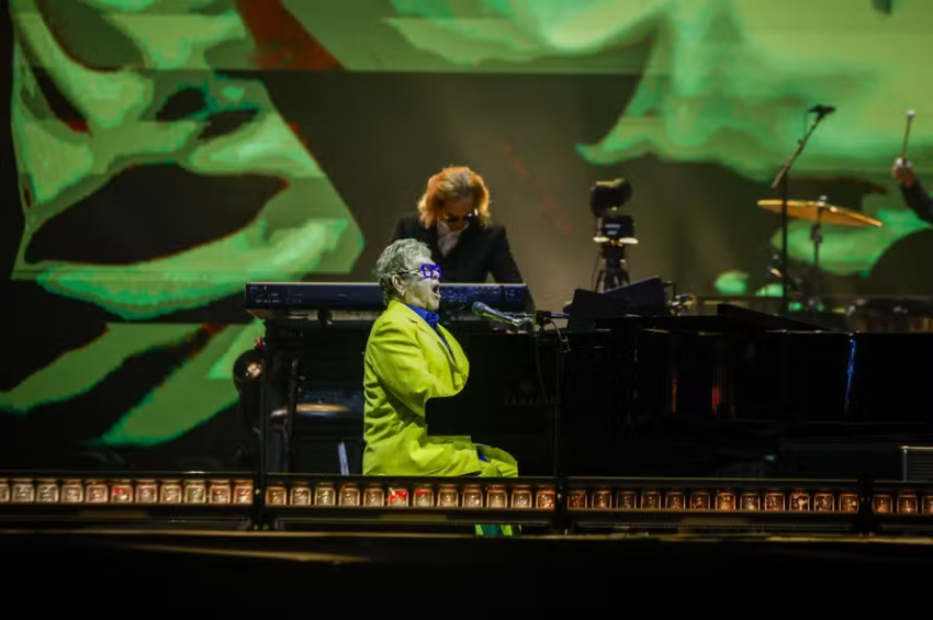 Elton John de terno amarelo tocando piano e cantando