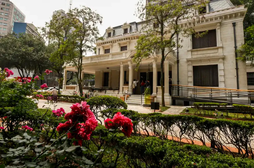 Um elegante casarão histórico de estilo neoclássico, cercado por jardins floridos e árvores em meio à paisagem urbana.