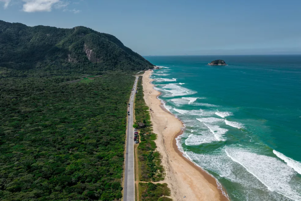 Uma extensa praia de areia clara acompanha o mar azul, cercada por montanhas e uma grande área de vegetação preservada.