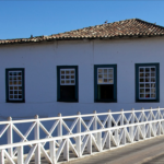 Fachada da Casa Velha da Ponte, na Cidade de Goiás, onde fica o Museu Casa de Cora Coralina: casarão colonial branco com janelas verdes, ao lado de uma ponte de madeira.